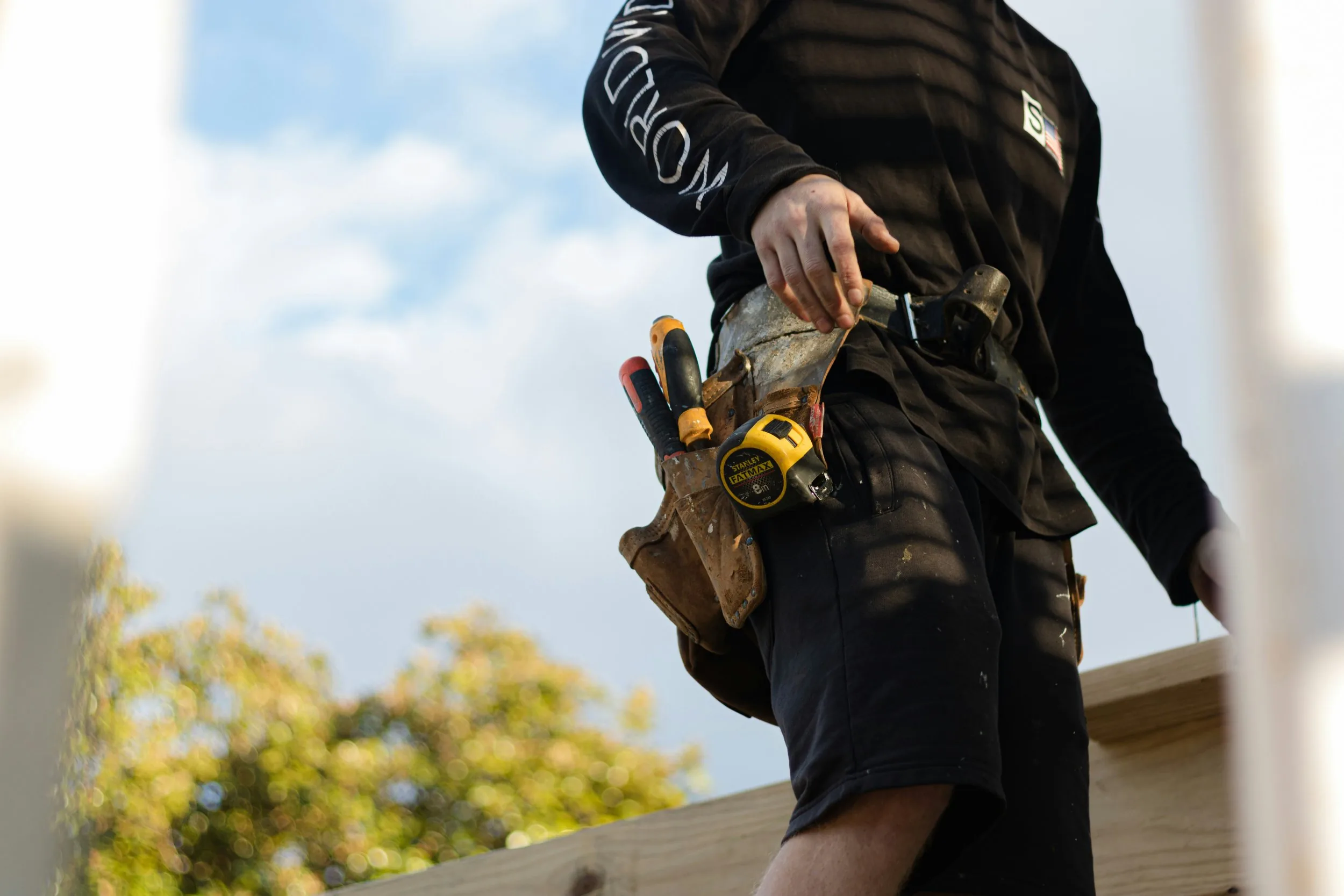 Builder in a black work shirt with a leather tool belt and tape measure, photographed from the chest down against the sky