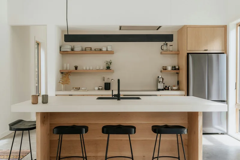 Wide view of a renovated kitchen with oak cabinets, a stainless fridge, white island bench and three black stools